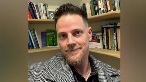 Adam Powell Dr Adam Powell, with short brown-reddish hair, with a ginger moustache and beard, wearing a black and white checked blazer, looks at the camera. A bookcase filled with books is behind him.
