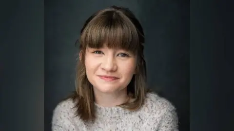 Yellow Belly Jade Croot, who has brown hair and is wearing a white jumper