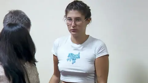 Rustavi 2 Bella Culley filmado no tribunal. Ela está usando óculos e tem cabelo castanho preso em uma trança e está vestindo uma blusa branca