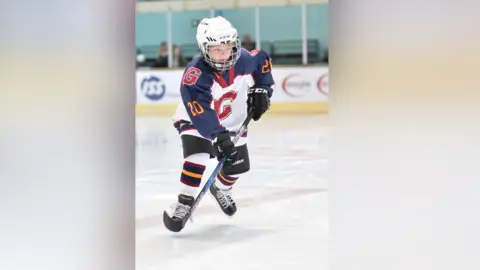 Guildford Junior Ice Hockey Club Rufus Dawson playing for Guildford back in 2019 at the age of eight. Rufus is skating on the ice and is wearing a white helmet, white, navy and red jersey with a 'G' in the middle, alongside gloves, kneepads and black skates. Rufus is holding his puck.