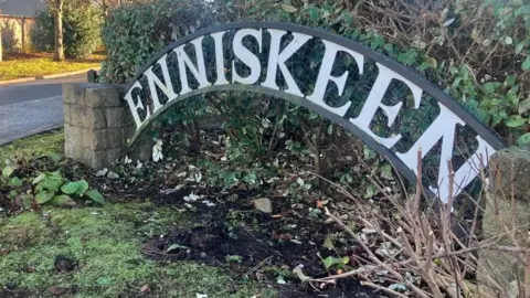 A sign displaying the name Enniskeen. It is in front of a green hedge and there are stone pillars either side
