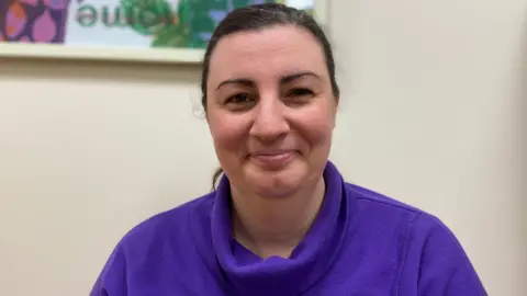 Stephanie McCarville wears a purple jumper. She has brown hair. There is artwork behind her on a white wall.