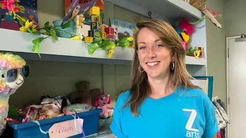 Elaine Doran/BBC Gilly Balfour with shoulder length brown hair wearing a blue t shirt with the logo Z arts on it smiling in front of shelves of colourful children's games and art materials