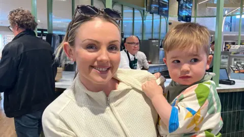 Judith is smiling at the camera wearing a white zip up and tortoise shell sunglasses on her hand. She has blond hair and blue eyes and is smiling at the camera. She has silver stud earrings in. She is holding her son who is blond with brown eyes. He is wearing a multi-coloured top and looking away from the camera. Behind them is a green and white counter with a waiter behind it.