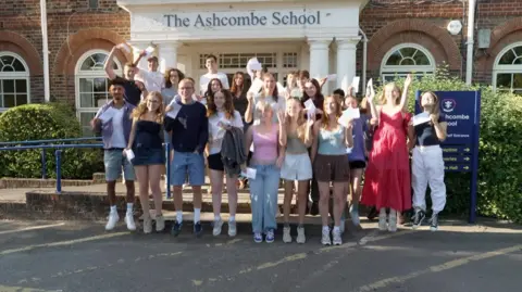 Students at The Ashcombe School in Dorking jumping for joy after receiving their results. They are all stood outside the school's entrance and are holding their results paper.