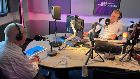 Lily-May Symonds/BBC Presenter Jonathan Vernon-Smith talking to Bedfordshire Police and Crime Commissioner John Tizard in a radio station studio. They are both wearing headphones and speaking across a desk, which has monitors on it.