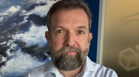 Who are the workers who will fire Scotland's space industry? 5 A man with grey hair and a grey beard looks into the camera wearing a neutral expression. He is wearing a white shirt and is standing in front of a photo of Earth taken from space.