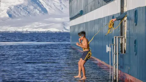 Yuam jumping into sub-zero water off a ship wearing blue swim shorts and goggles has a yellow rope tied around him for safety.