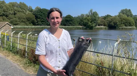 Jane Pearce holds a scale model of the phosphate removal tool in front of Chew Valley lake on a sunny day. She wears a white shirt and shorts. The model consists of black, charcoal-like pellets in a black mesh bag. 