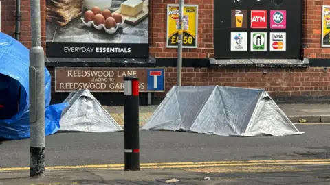 Two silver small tents and blue tarpaulin over a car next to the Reedswood Lane sign