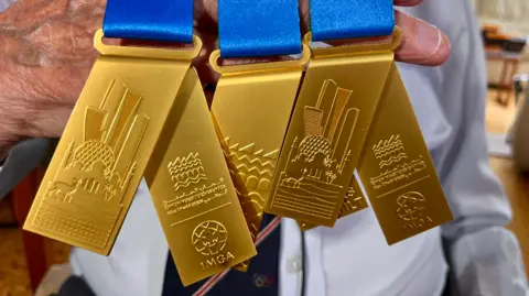 Martin Giles/BBC A close-up image of five gold medals draped over the older-looking hand of a man who is wearing a pale blue shirt with a navy and red-stripped tie. 