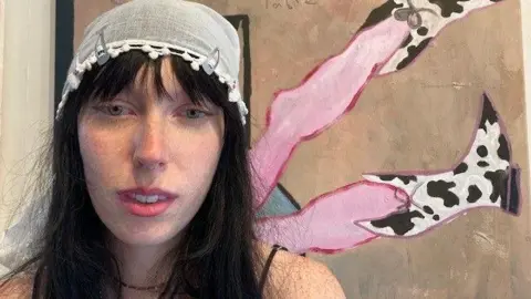 BBC Poppy Baynham, uma mulher com longos cabelos pretos e um guardanapo de renda na cabeça, em pé em frente a uma de suas pinturas