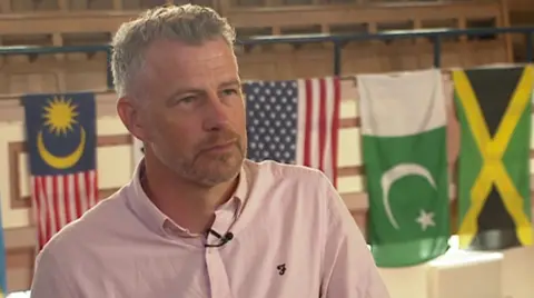 Ben Gibbins with salt and pepper hair sporting a pink shirt and a three-day stubble. In the background are several flags mounted to the wall, including Jamaica's flag.
