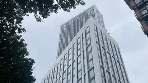 A view looking up at Birmingham's One Eastside from street level. The tall white and grey tower rises into a cloudy sky, with a shorter block to its right and green tree branches framing the top of the image.