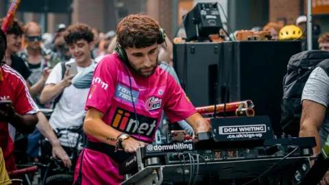 Dom Whiting Dom Whiting on one of his bike raves, surrounded by people riding bikes. He is playing music on his decks.