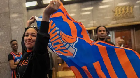 A Knicks fan in front of a building waves an orange and blue flag