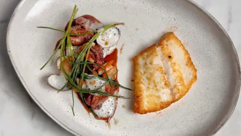 BBC Food A close-up of a dish showing a pan-fried fish steak and accompanying salad that includes cockles, tomatoes and samphire. The food is presented on a grey ceramic plate. 