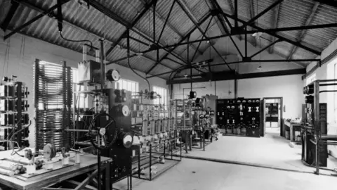 A transmitter hall shows various equipment bays with valves and other components in racking. There are several dials fixed to the racks. There is a thick wire running across the floor. There is a metal grid supporting the grey roof. Windows are visible to the left.