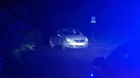 West Midlands Police A silver car parked on a rural roadside at night, its registration blurred.