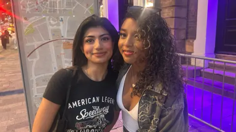 Two young women are smiling. On the left, the young woman has long black hair and a black t-shirt with white writing. On the left, the young woman has curly brown hair, sunglasses on her head, a white top and denim jacket