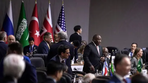 Getty Men in women in formal wear stand up from their seats in a hall against a backdrop of national flags.   