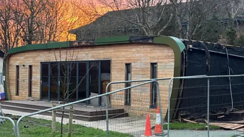 BBC/Paul O'Gorman A single-storey wooden building with a damaged roof and side. It has four slim black-framed windows and a large multi-door opening in the middle.