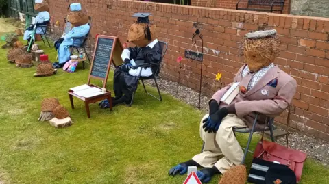 Nigel Taylor Four scarecrows resembling hedgehogs positioned in chairs in a row. Each character is distinctly dressed in suits. They are against a brick wall.