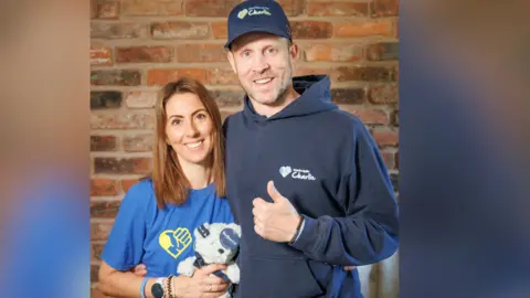 Robinson family/ThumbsUpForCharlie Couple wearing blue t-shirts. A woman with brown hair stands on the left and holds a teddy bear - a man stands on the right with a thumb up. 