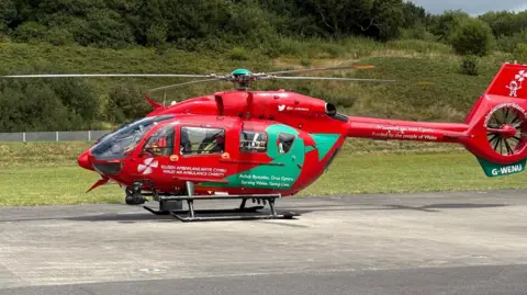 BBC The red Wales Air Ambulance helicopter is on a tarmac surface. It has the name of the charity in white writing on the side of the helicopter, as well as the slogan "serving Wales - saving lives" and "funded by the people of Wales". It also has the green loop of the Welsh dragon's tail painted on it. There are crew members visible inside the aircraft as it prepares for take-off.