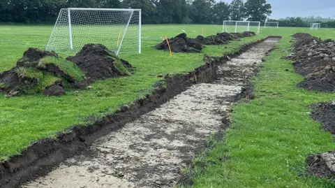 A number of football pitches with goalposts. A long trench has been dug in the grass with piles of dirt piled on either side. There are trees and hedges in the background.