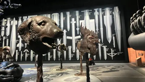 Photograph of the Ai Weiwei exhibition at Manchester's Aviva Studios. The image shows animal head sculptures and a large wall mural.
