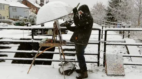 Rob Pointon A man paints by a lock in heavy snow. His easel is covered by an umbrella. He is standing by a fence and houses.