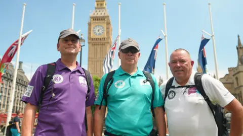 The three dads standing in a line in front of Big Ben. Tim Owen is wearing a purple polo shirt and a grey cap and smiling into the camera. Mike Palmer is standing next to him with a turquoise polo shirt and grey cap and black sunglasses. Andy Airey is on the right and is wearing a white polo shirt. He has a shaved head and is smiling into the camera.