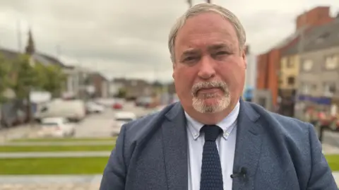 Colm Gildernew is a big man with short grey hair and a grey goatee. He is wearing a grey-blue suit. 