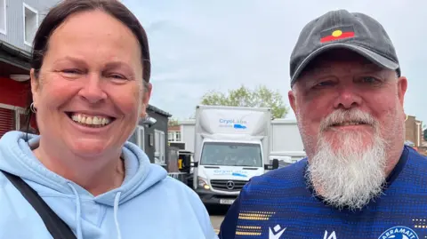 Tracy and Shane Lester smiling at the camera. Shane wears a cap and a football shirt and has a long white goatee. Tracy has her dark hair tied back and is wearing a light blue hoodie.