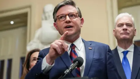 Speaker Mike Johnson in a blue suit, raising his hand while talking into a microphone