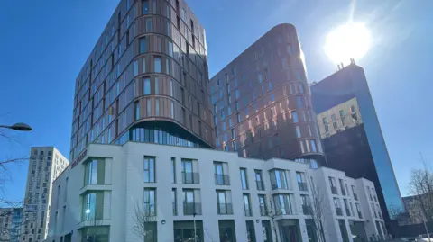 The university's housing building, gray with rectangular windows and a brown building with two curves on top of it. The background is of blue sky and sun.