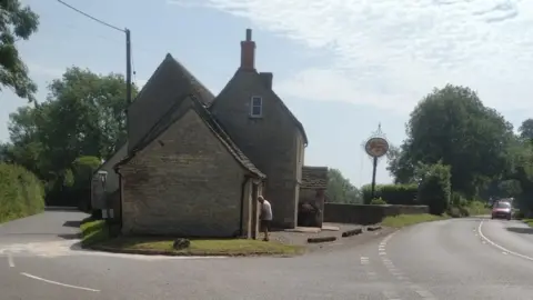 A stone-brick pub stands at the intersection of two roads. There are lots of trees and hedgerows around, and it is a sunny day with light cloud cover. 