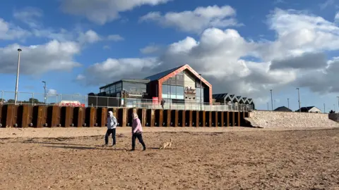 Steel piling have been put in place on the sea wall as part of phase one of the project. The large steel piling sits below the Mickey's Restaurant establishment which overlooks Exmouth beach. Two people are walking along the sand. A dog is at their heels.