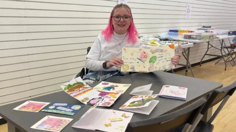 Katie Callaghan Katie Callaghan is wearing a white roll neck and has pink hair. She is sat at a grey table which is covered in handmade cards. She is holding one open towards the camera.