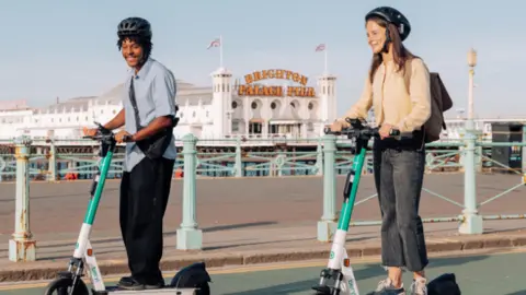 Two people on e-scooters on a road. They are wearing helmets. A pier can be seen in the background.