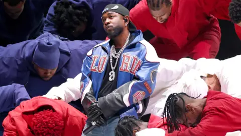 Getty Images Kendrick Lamar during his Super Bowl halftime show. Kendrick poses with his arms folded across his front, holding a mic. He wears a blue leather jacket over a black T-shirt with blue flared jeans, a black cap twisted back and a large diamond necklace with a lower case 'a'. Dancers dressed in the red, white and blue of the US flag surround him as he looks up into the crowd. 