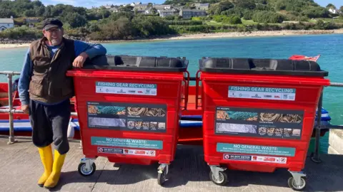 Duchy of Cornwall A fisherman smiling at he stands next to two large rectangular recycling bins which have labels which say: FISHING GEAR RECYCLING and NO GENERAL WASTE with pictures of fishing gear. Behind him is a bay and shoreline.