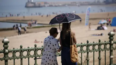 EPA Two women with an umbrella walk past a green fence. A beach with lots of people on it is visible in the background.