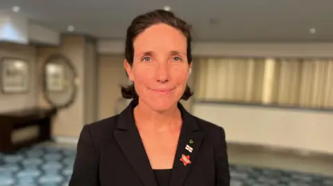 BBC Lindsay de Sausmarez looking at the camera, she is a woman with close black hair, wearing a black jacket with three pin badges on the lapel.