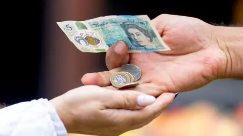 Two hands with a £5 note and coins being passed over.