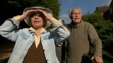 Channel 4 Prunella Scales and Timothy West on a canal boat. Timothy is in brown jumper and looking off in the distance. Prunella is in a yellow shirt, brown waistcoat with a blue denim jacket. She is also looking in to the distance but shielding her eyes from the sun.
