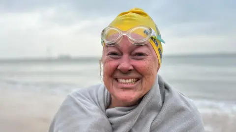 Helen Jenkins smiles into the camera after a swim, wrapped in a towel and wearing a yellow swimming hat, with goggles pushed up on the top of her head.