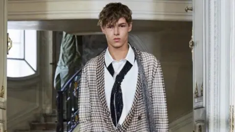 Guernsey sixth form student Jack Paulsen modelling for fashion brand Dior in Paris. He is looking at the camera and is wearing a jacket, an embroidered tie and a very thin scarf over his shoulder. 

