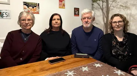 Four people sitting around a wooden table. On the left is a woman wearing a red cardigan and has grey hair and glasses, she is frowning. Next to her on the right is a woman with medium length brown hair and a black top, she is also frowning. On her right is a man wearing a blue shirt and has grey hair. He is frowning too. Then, next to him is a woman wearing a black cardigan and Christmas themed dress, she has blonde hair and is smiling.